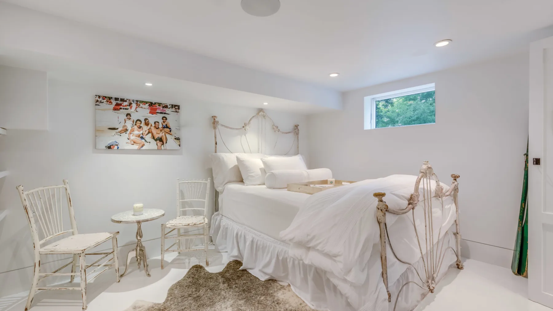 A bright bedroom with a white bed, pale furnishings, and a small wall picture.