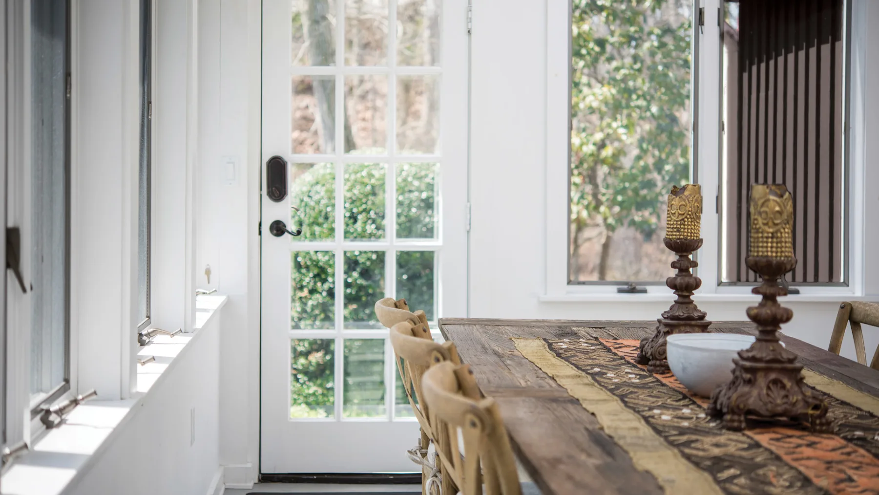 A bright dining area with a long wooden table beside tall windows and a glass-paneled door.