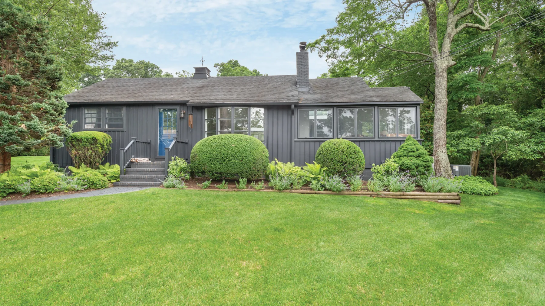 A dark single-story house stands behind a broad green lawn and garden shrubs.