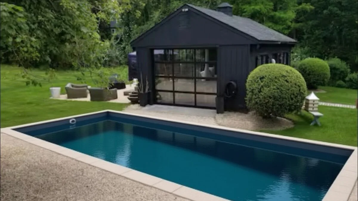 An outdoor swimming pool sits beside a dark pavilion, with trees and shrubs around the yard.