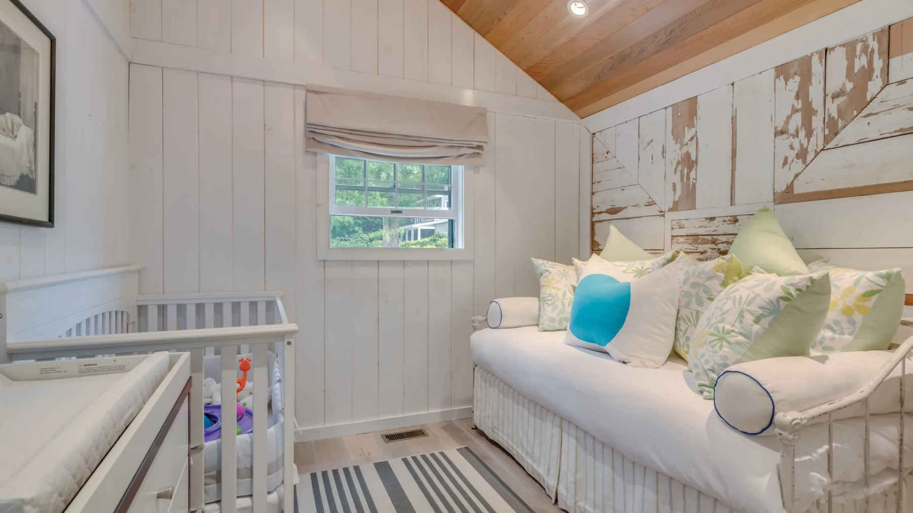 A bright bedroom with a white bed, striped rug, and crib beside the window.