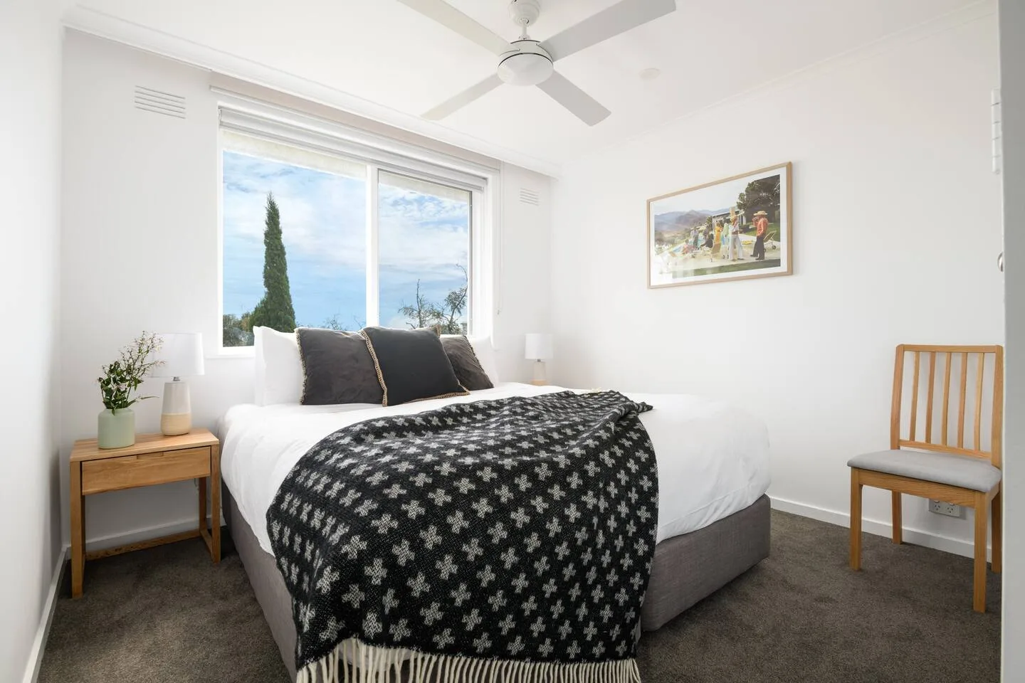 A bright bedroom has a bed with dark patterned bedding and a chair by the wall.