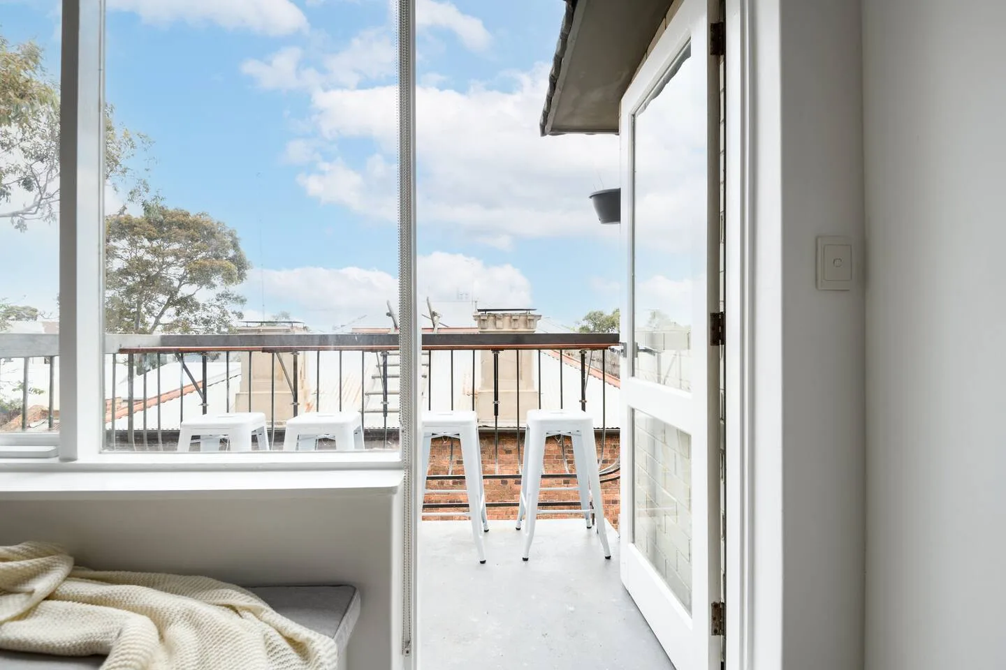 Una habitación luminosa da a un balcón con asientos de barra y vistas a los tejados cercanos.