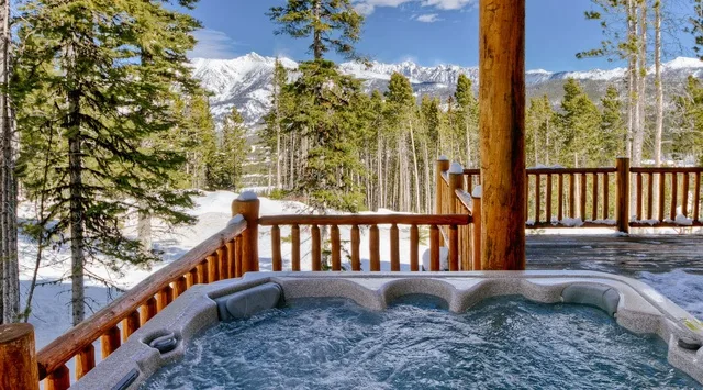 An outdoor hot tub sits beside a wooden deck, with snowy mountains beyond the trees.