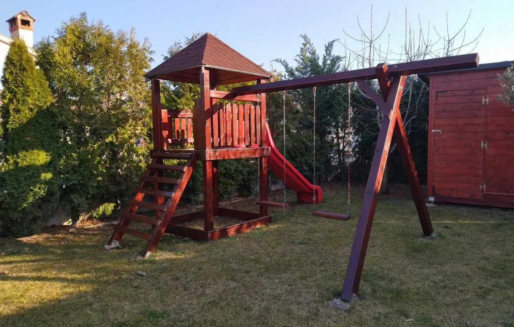Una estructura de juegos de madera con tobogán y columpio está en un patio con césped.