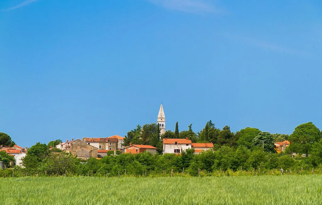 Un pueblo con un campanario se alza tras un campo verde bajo un cielo despejado.