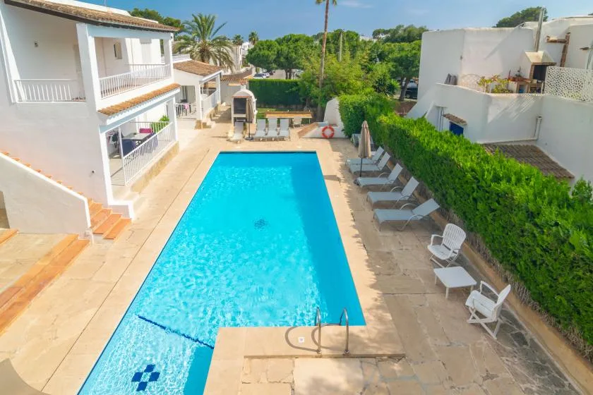 A swimming pool sits between white buildings, with lounge seating along the paved deck.