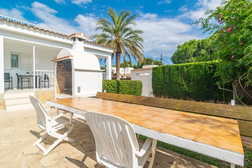 A sunny outdoor dining patio with a long table beside a white house and garden hedge.