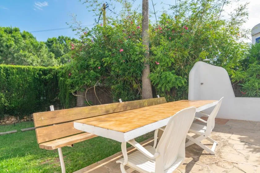 An outdoor dining area with a wooden table and white seating beside a leafy hedge.