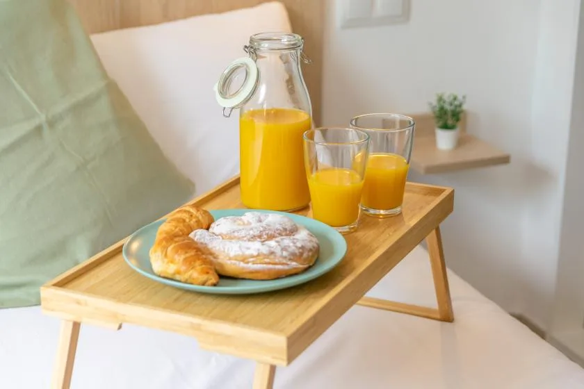 Breakfast with pastries and orange juice rests on a wooden tray beside a bed.