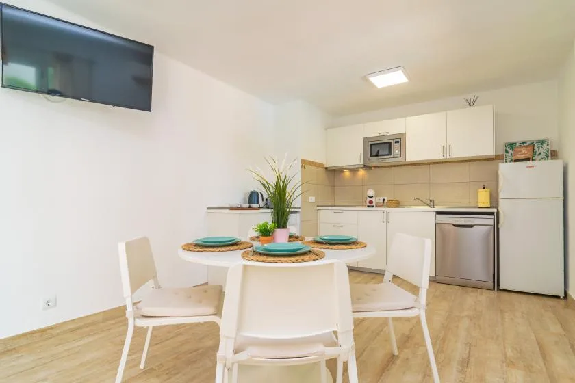 A bright dining area with a round table and a compact kitchen behind it.