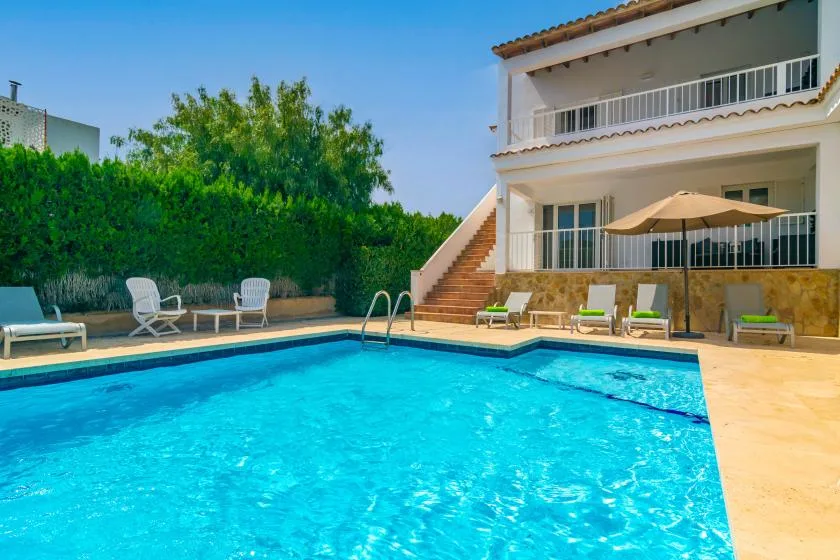 Una piscina exterior luminosa está junto a una casa blanca de varias plantas y asientos en el patio.