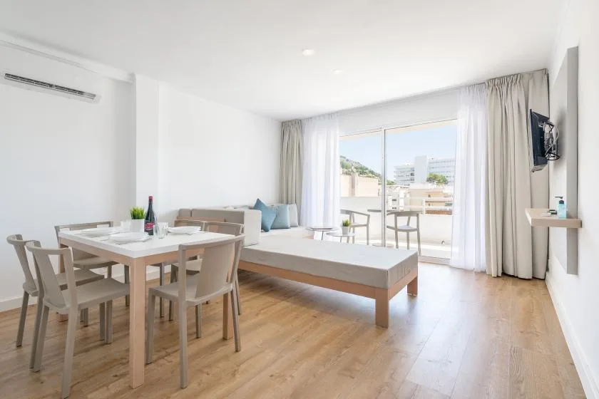 A bright room combines a dining table with seating and a bed beside a large window.