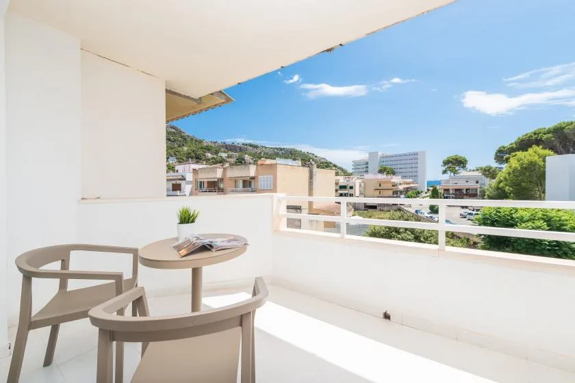 A bright balcony with outdoor seating overlooks nearby buildings and a green hillside.