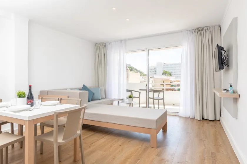 A bright studio room has a bed, a dining table, and glass doors opening onto a balcony.