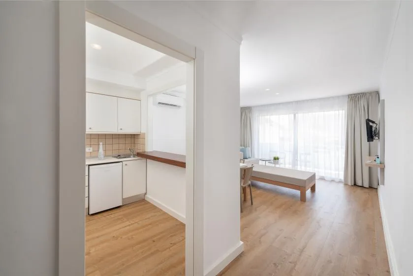 A bright open-plan living area and kitchenette with wood flooring and large curtained windows.
