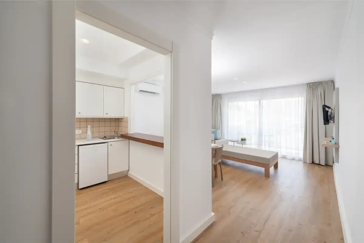 A bright living room with a small kitchen area and large curtained windows.