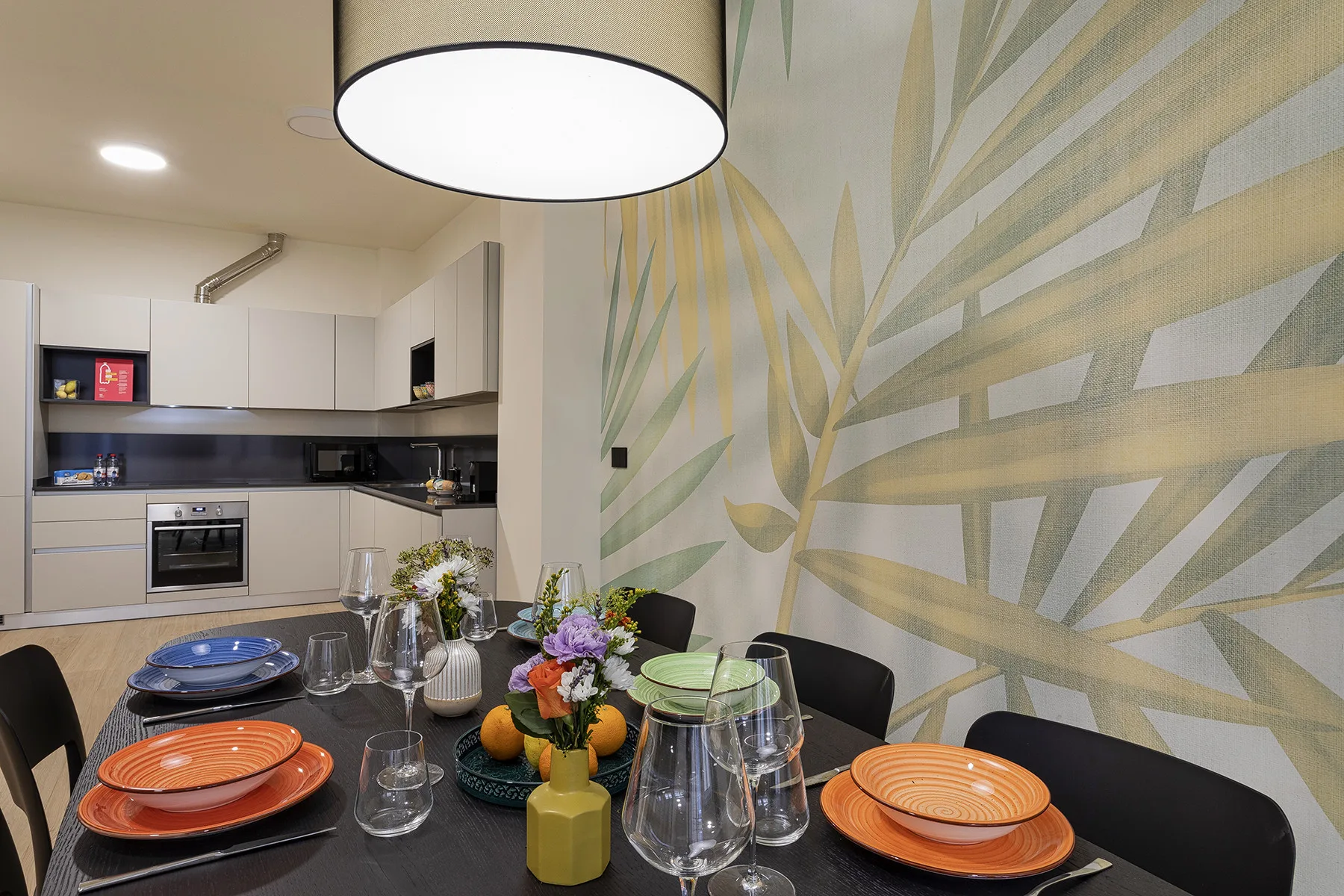 A dining table set with dishes and flowers sits in front of a mural, with a kitchen beyond.
