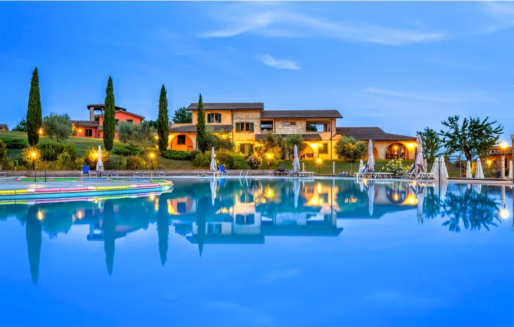A swimming pool reflects a row of illuminated buildings beneath a blue evening sky.
