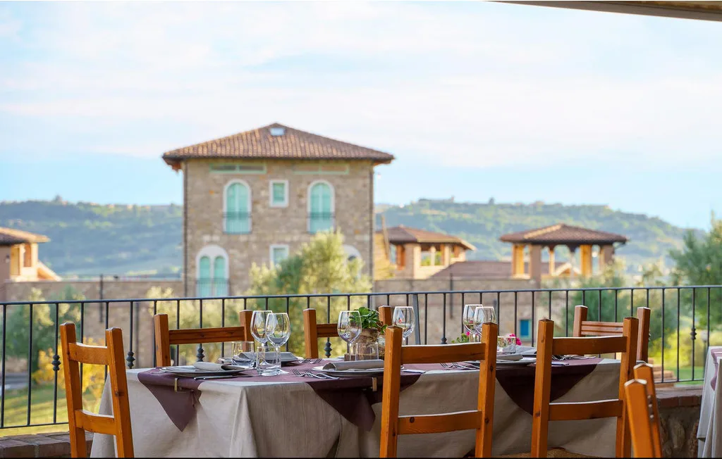 An outdoor dining table is set beside a railing, with hills and buildings beyond.