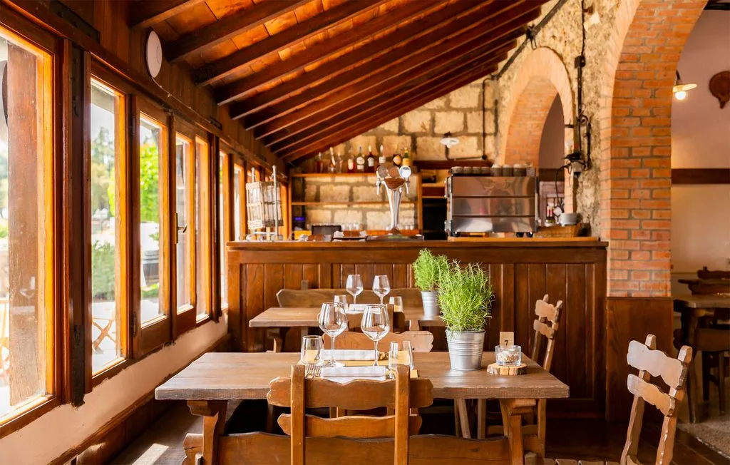 A rustic dining room with wooden tables, tall windows, and a bar.