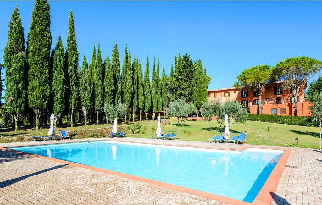 An outdoor swimming pool sits beside a grassy lawn with tall trees and a building.
