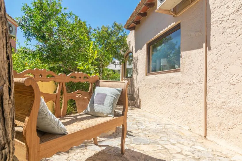 A sunny stone patio with wooden seating and leafy trees.