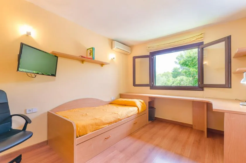 A bedroom with a single bed, desk, and open window overlooking greenery.