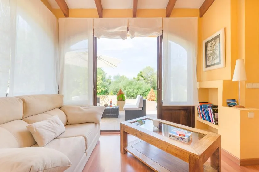 A bright living room with seating, a coffee table, and large windows opening to greenery.