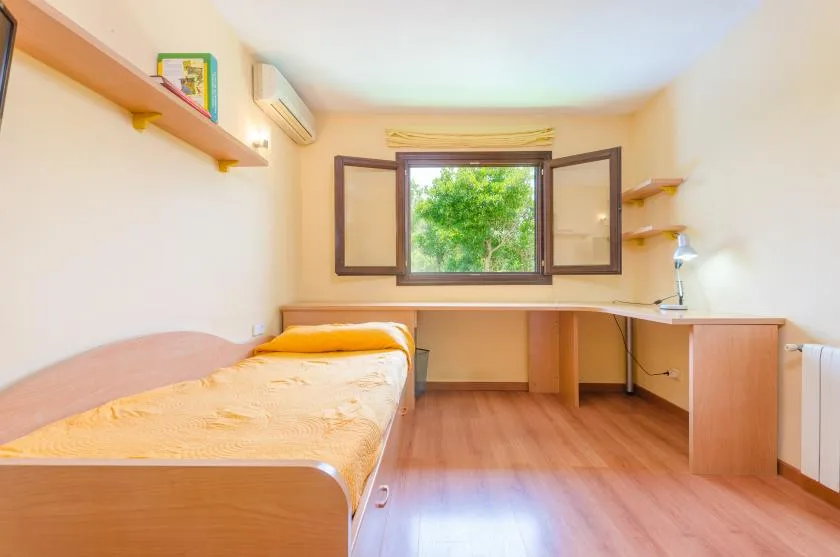 A bedroom with a single bed, a desk, and an open window overlooking greenery.