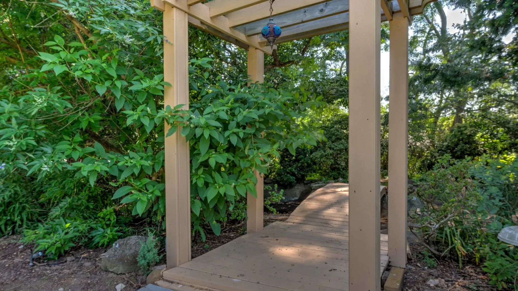 A wooden garden walkway passes through dense greenery beneath a simple overhead structure.
