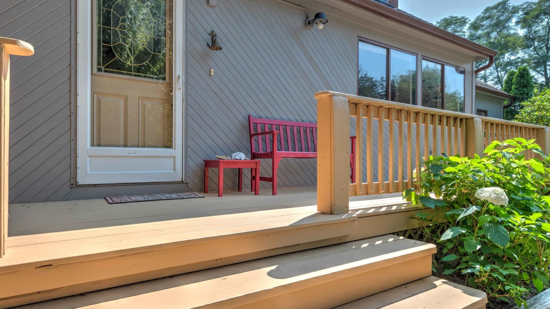 A wooden deck with steps, a red bench, and railing beside a house.
