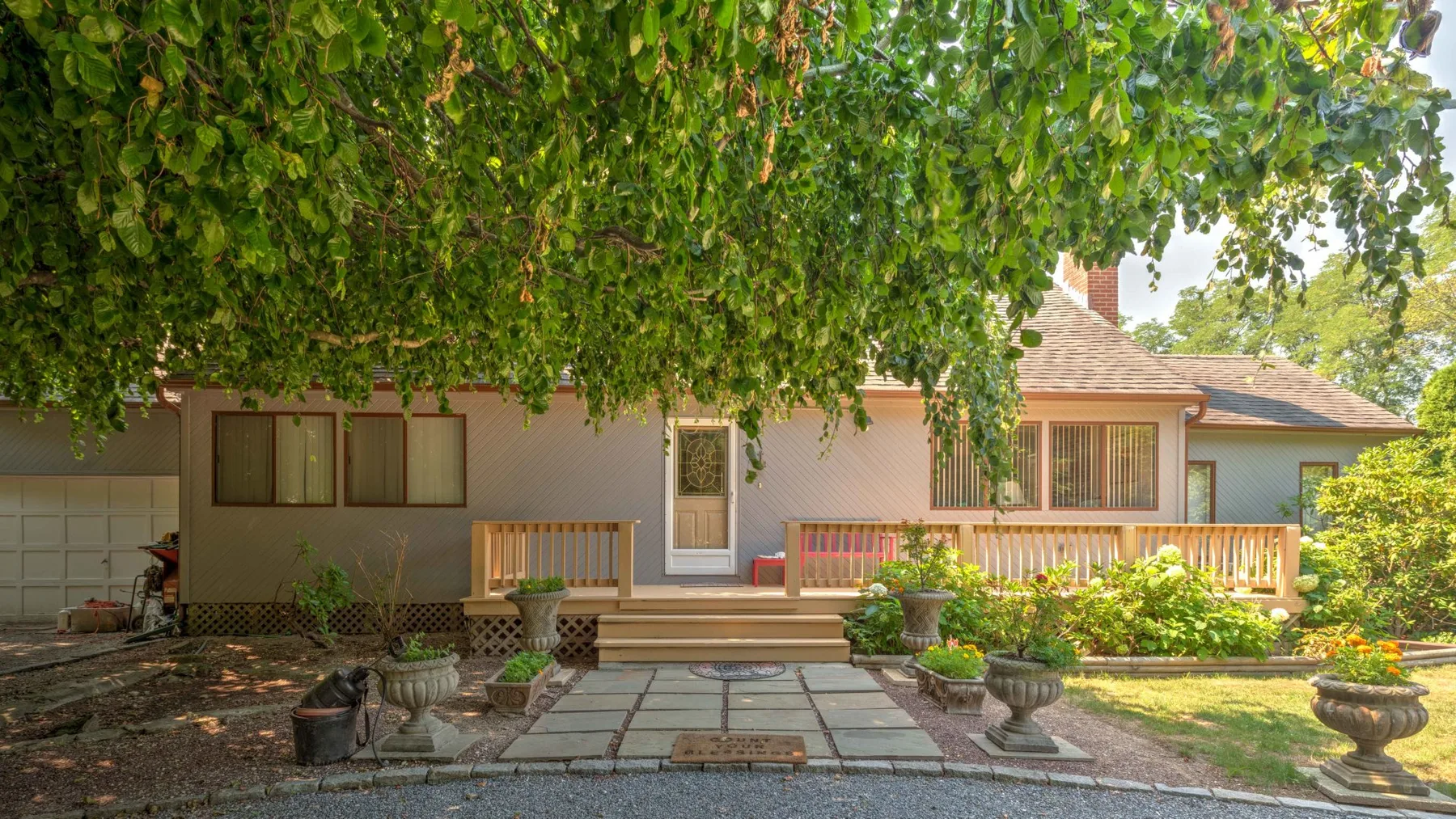 A house exterior with a paved patio, wooden deck, and leafy tree canopy.