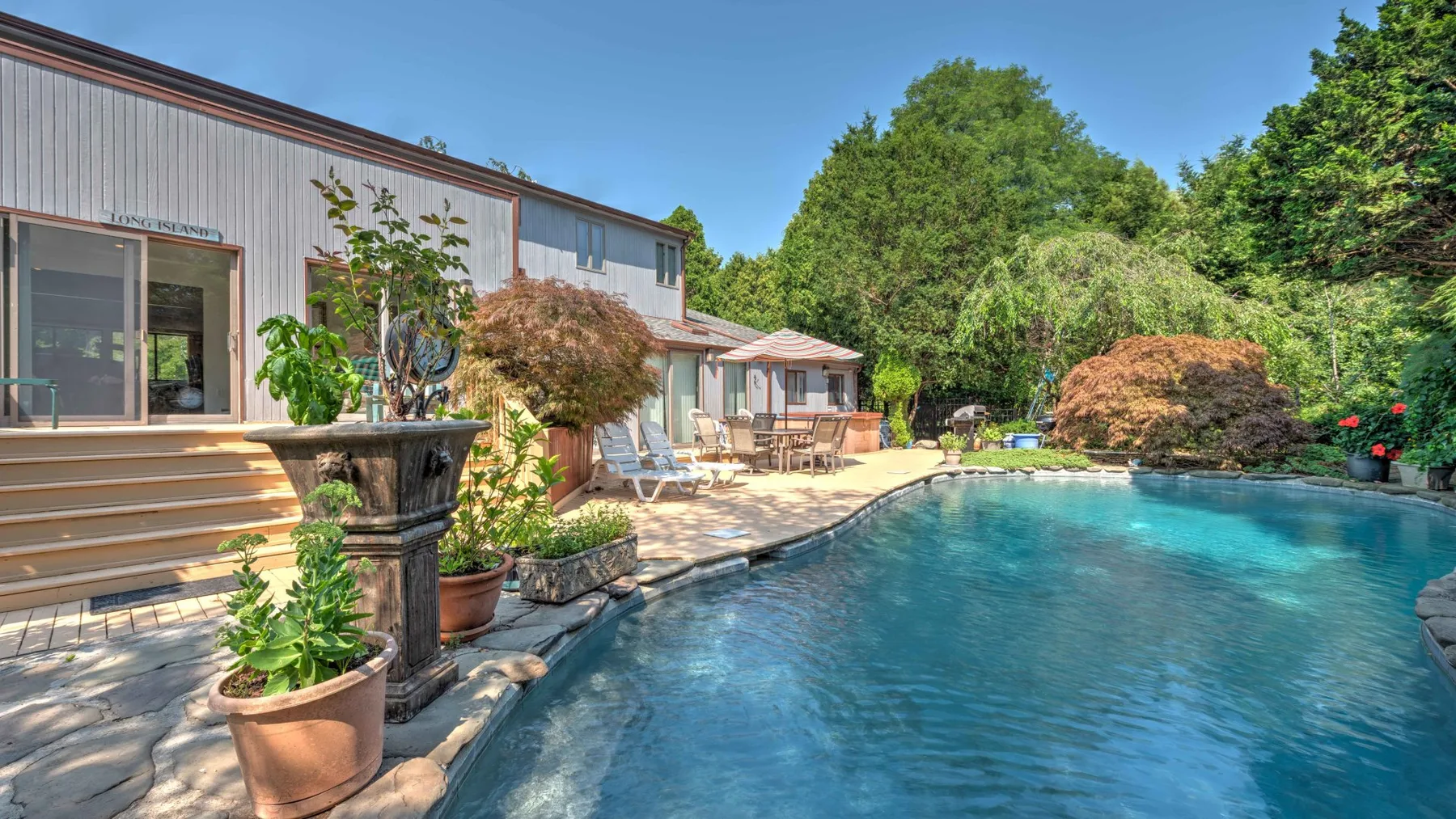 A swimming pool beside a house, patio seating, and landscaped greenery.