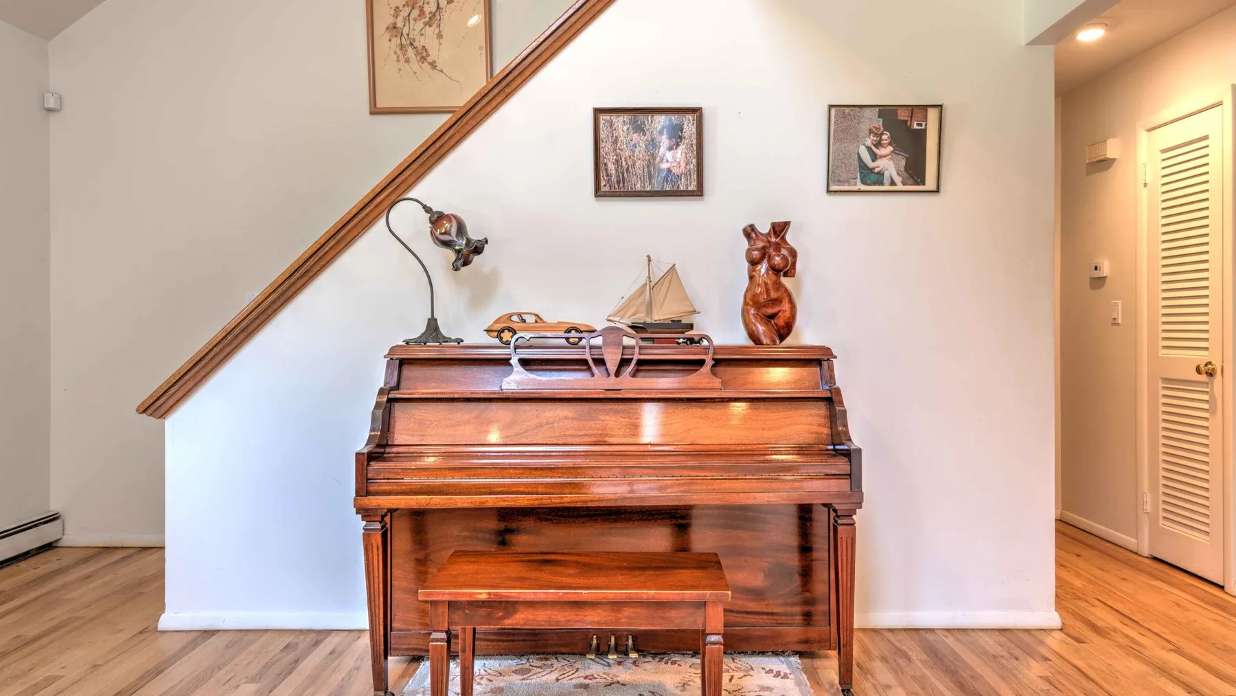 A wooden piano and bench sit beneath a staircase in a bright room.