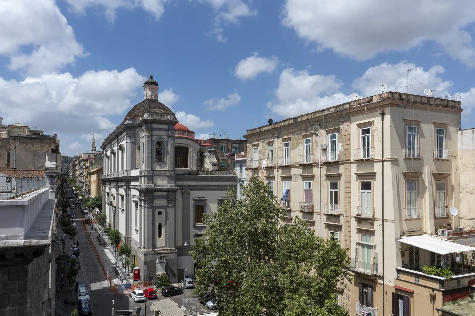 Una calle urbana flanqueada por edificios históricos, con una iglesia abovedada y árboles frondosos.