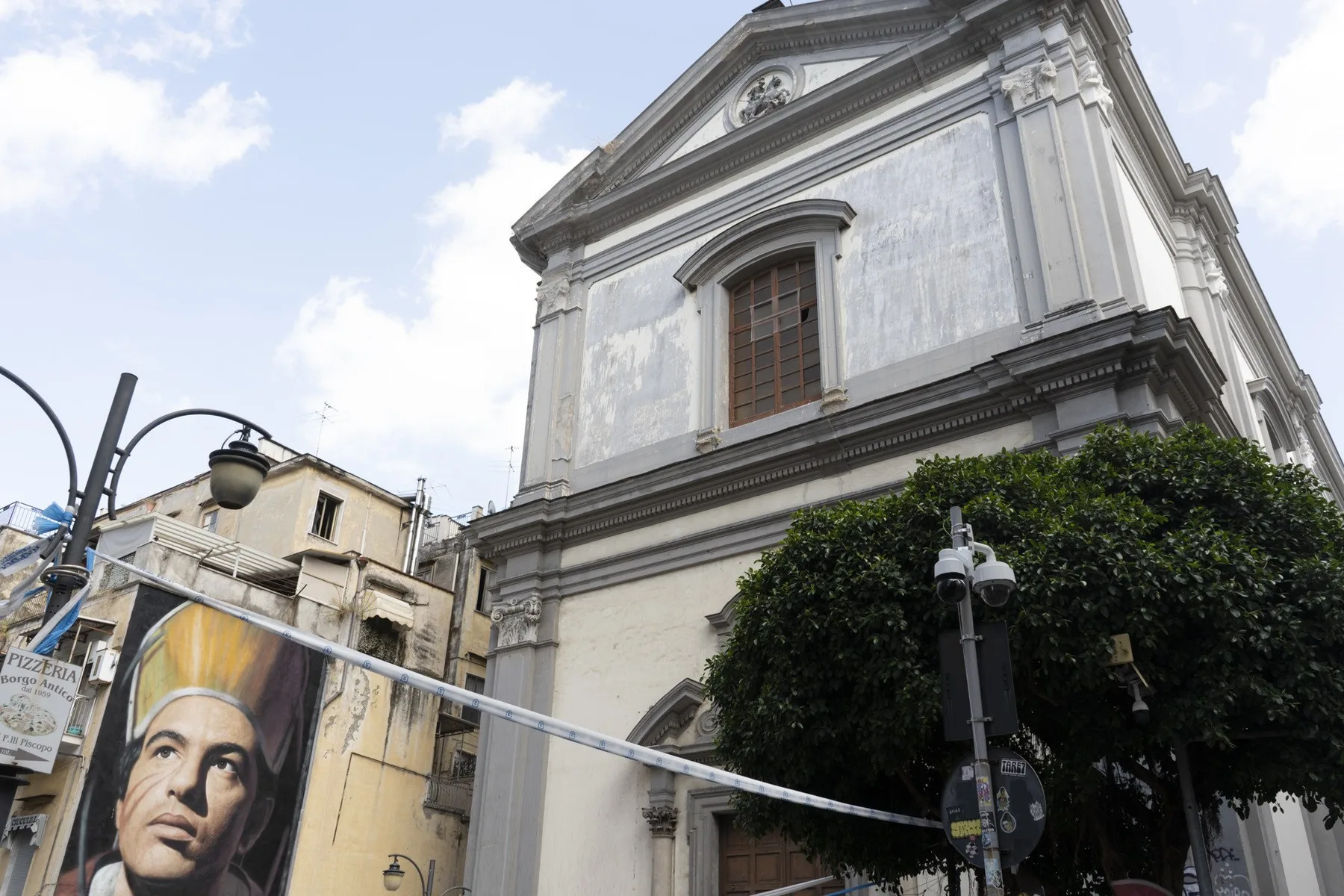 La fachada de una gran iglesia blanca se alza sobre la calle, con un árbol frondoso delante.