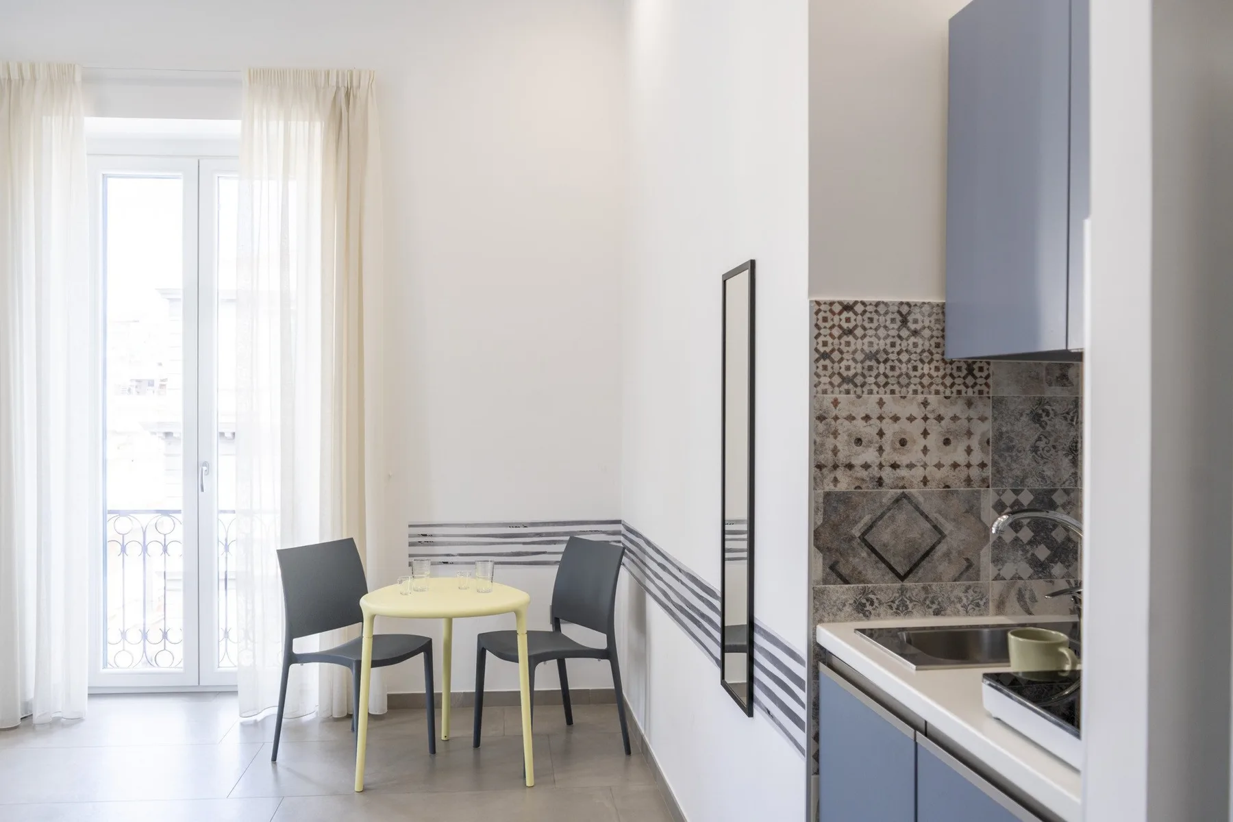 Un comedor luminoso con una mesa pequeña junto a ventanas altas y una cocina compacta.