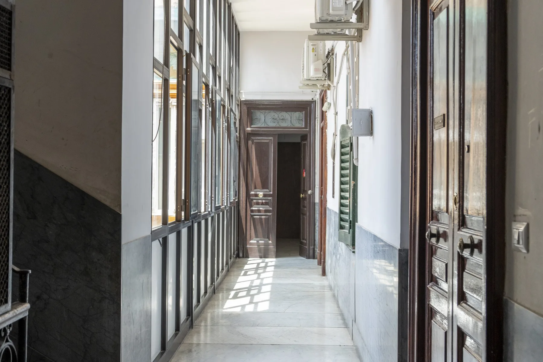 Un pasillo interior estrecho con ventanas altas a un lado y puertas al otro.