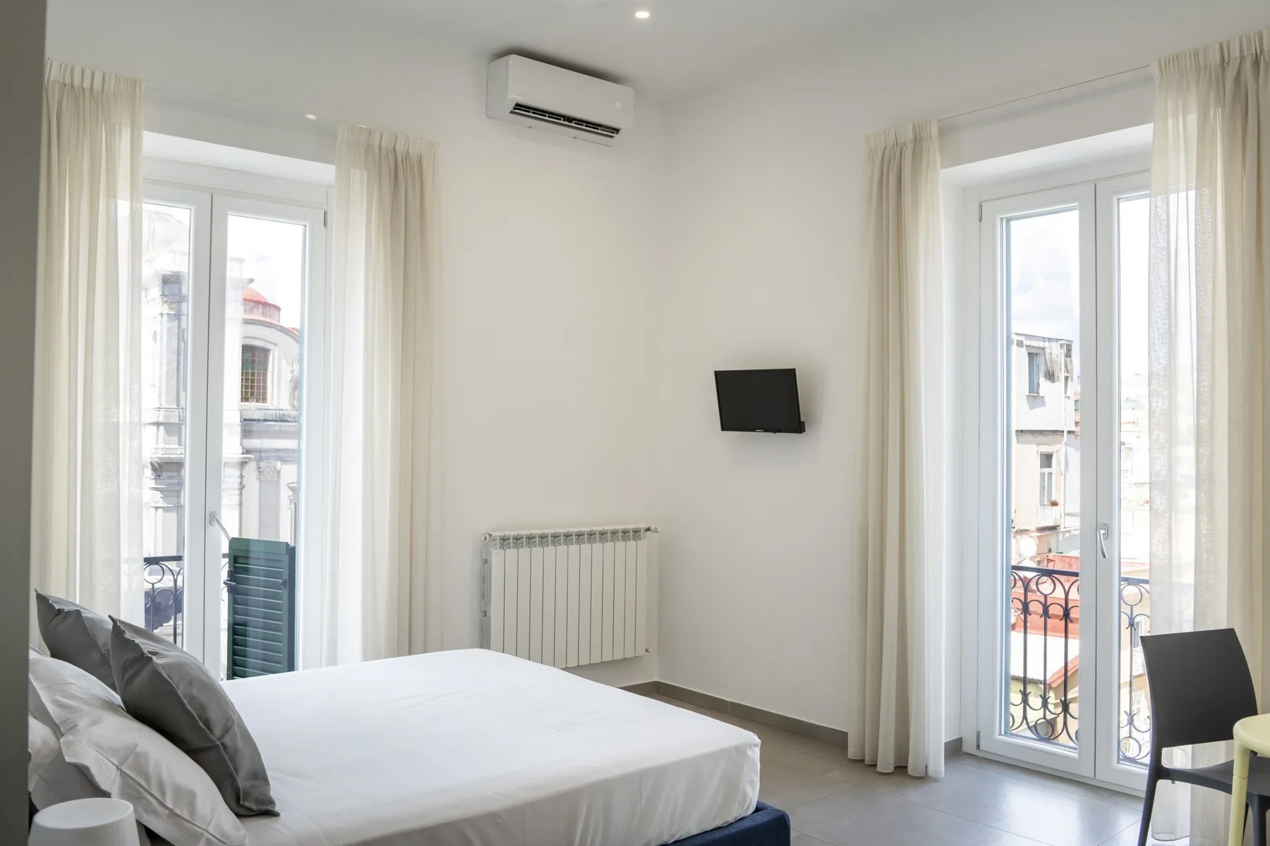 Dormitorio luminoso con una cama, dos ventanas altas y un televisor montado en la pared.