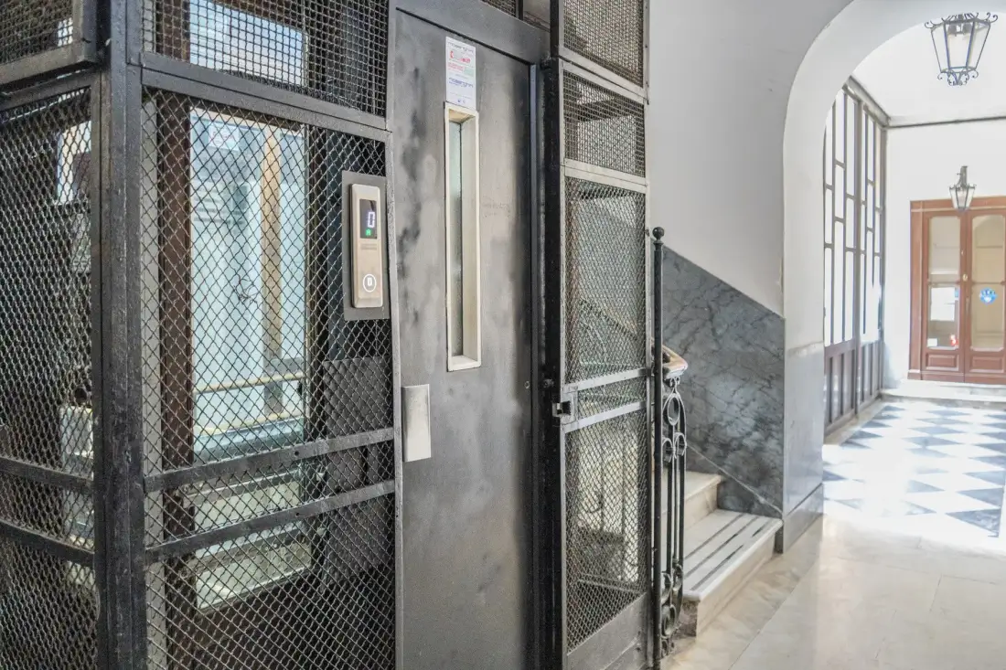 Un vestíbulo interior con un ascensor de malla metálica y un pasillo embaldosado.