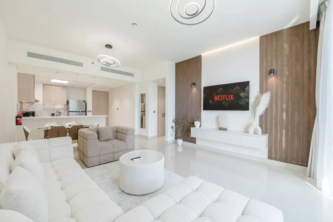 Una sala de estar luminosa con asientos blancos y un televisor, abierta a una cocina.