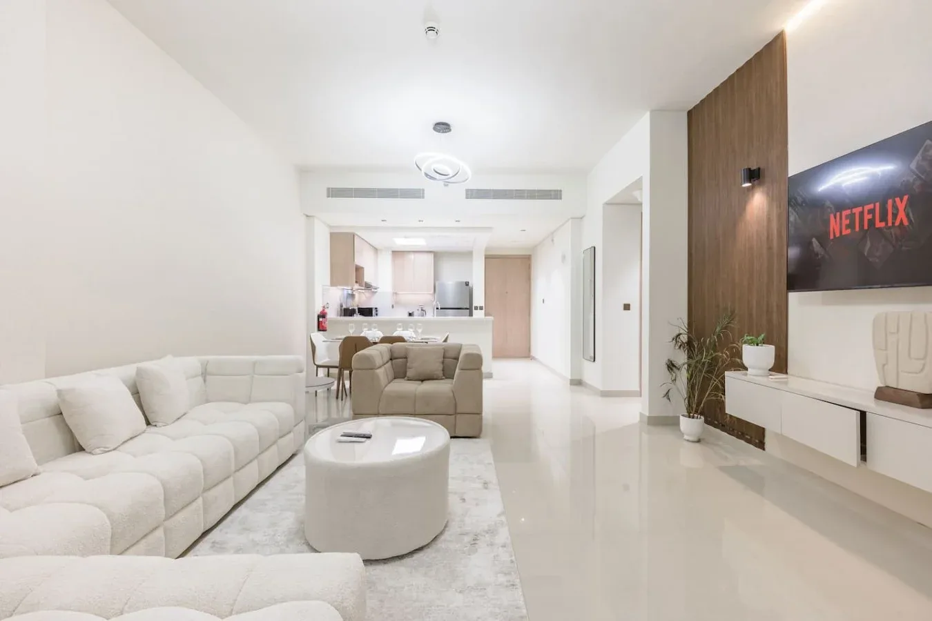 Una sala luminosa con asientos claros, un televisor y una cocina abierta al fondo.