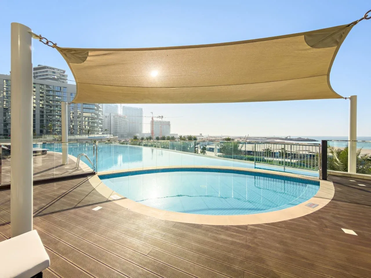 Una piscina al aire libre está junto a una terraza de madera bajo una lona de sombra.