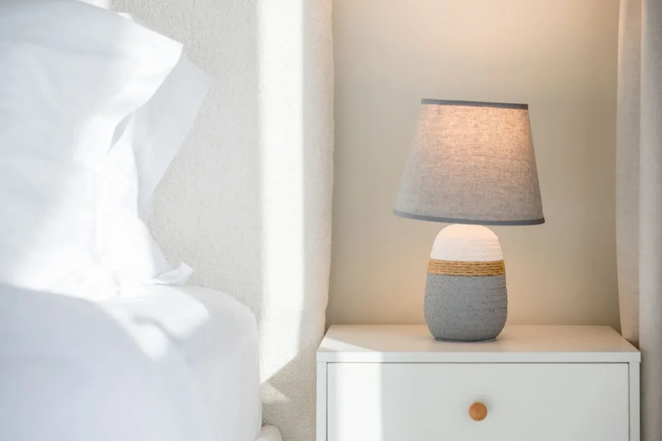 Una mesita de noche sostiene una lámpara junto a ropa de cama blanca.