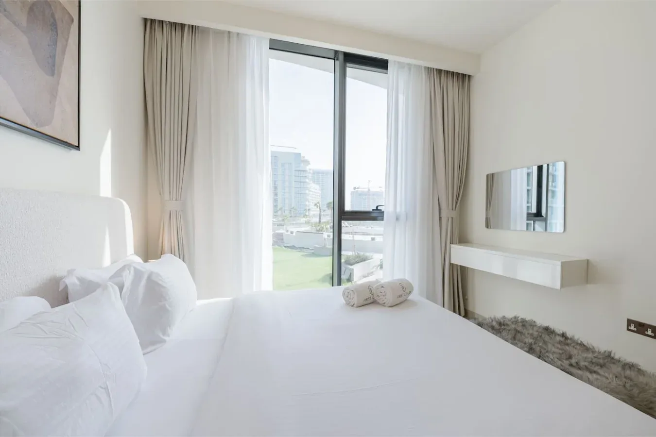 Un dormitorio luminoso con ropa de cama blanca y una ventana grande con vistas a edificios cercanos.