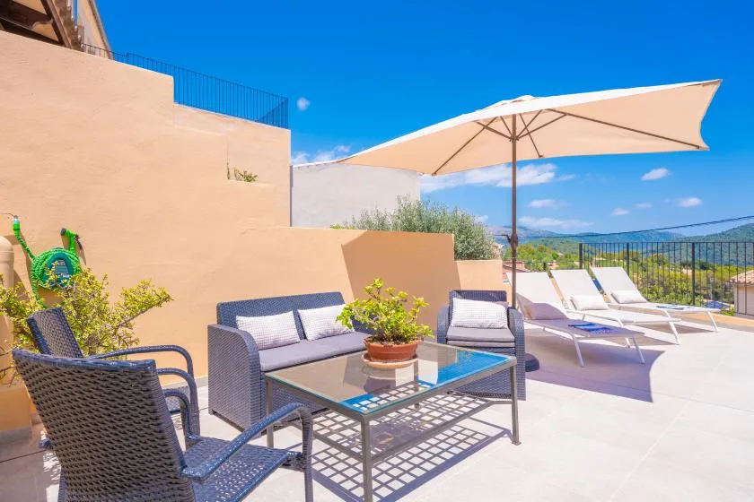 Un patio exterior tiene asientos, una mesa y tumbonas bajo una sombrilla grande.