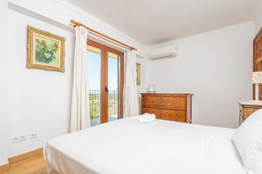 Un dormitorio luminoso con una cama blanca, una cómoda de madera y puertas acristaladas al balcón.