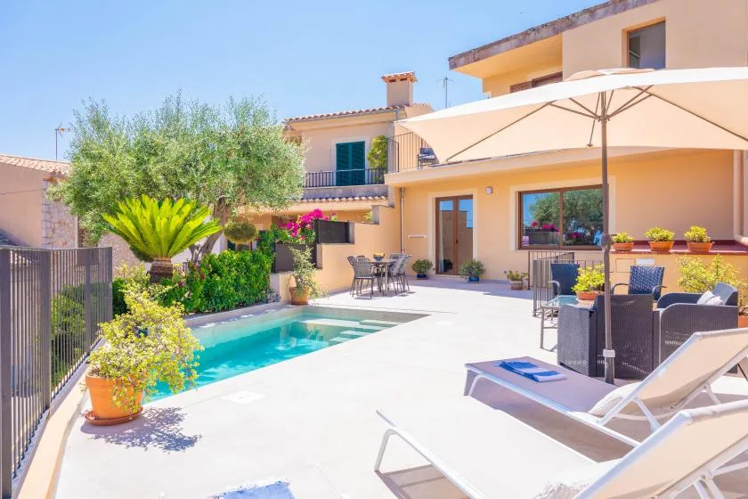 Un patio soleado con piscina, asientos al aire libre, sombrillas y plantas junto a la casa.