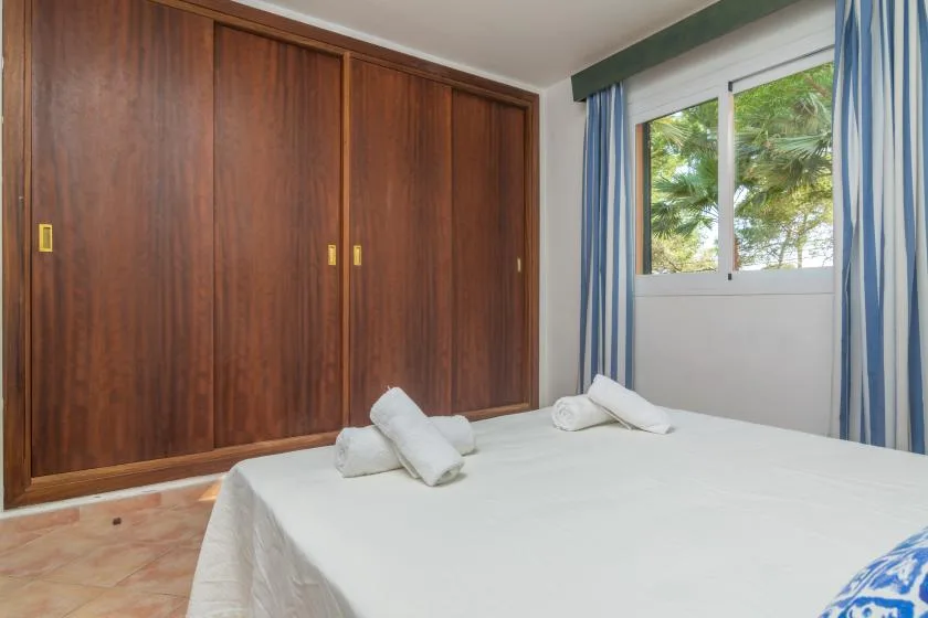 A bedroom with a white bed, dark wooden wardrobes, and a window overlooking greenery.